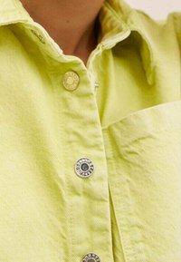 Camicia a maniche lunghe verde chiaro realizzata in tessuto testurizzato, con bottoni argentati e una tasca a toppa sul lato sinistro.