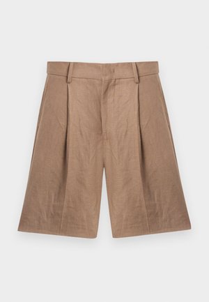 Homme debout les mains dans les poches, portant une chemise bleue à manches courtes, un short beige au genou et des sandales noires sur un fond uni.