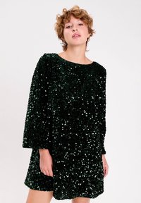 Une personne jeune avec des cheveux bouclés courts porte une robe ample en sequins vert foncé, se tenant devant un fond uni clair.