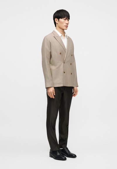 Μπεζ διπλόσαρκο σακάκι με καφέ κουμπιά, συνδυασμένο με καφέ παντελόνι με σχέδια και μαύρα δερμάτινα παπούτσια. Λευκό πουκάμισο με γιακά από κάτω.