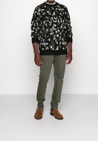 Pull noir avec un motif abstrait blanc et vert, associé à un pantalon vert olive et des bottines en daim marron.