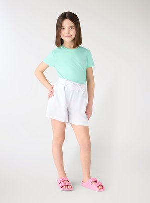 Ragazza giovane in piedi con la mano sui fianchi, indossa una maglietta verde menta, pantaloncini bianchi e sandali rosa su uno sfondo chiaro uniforme.