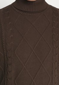 Pull à col roulé marron en maille avec motifs losanges et torsades sur le devant et les manches.