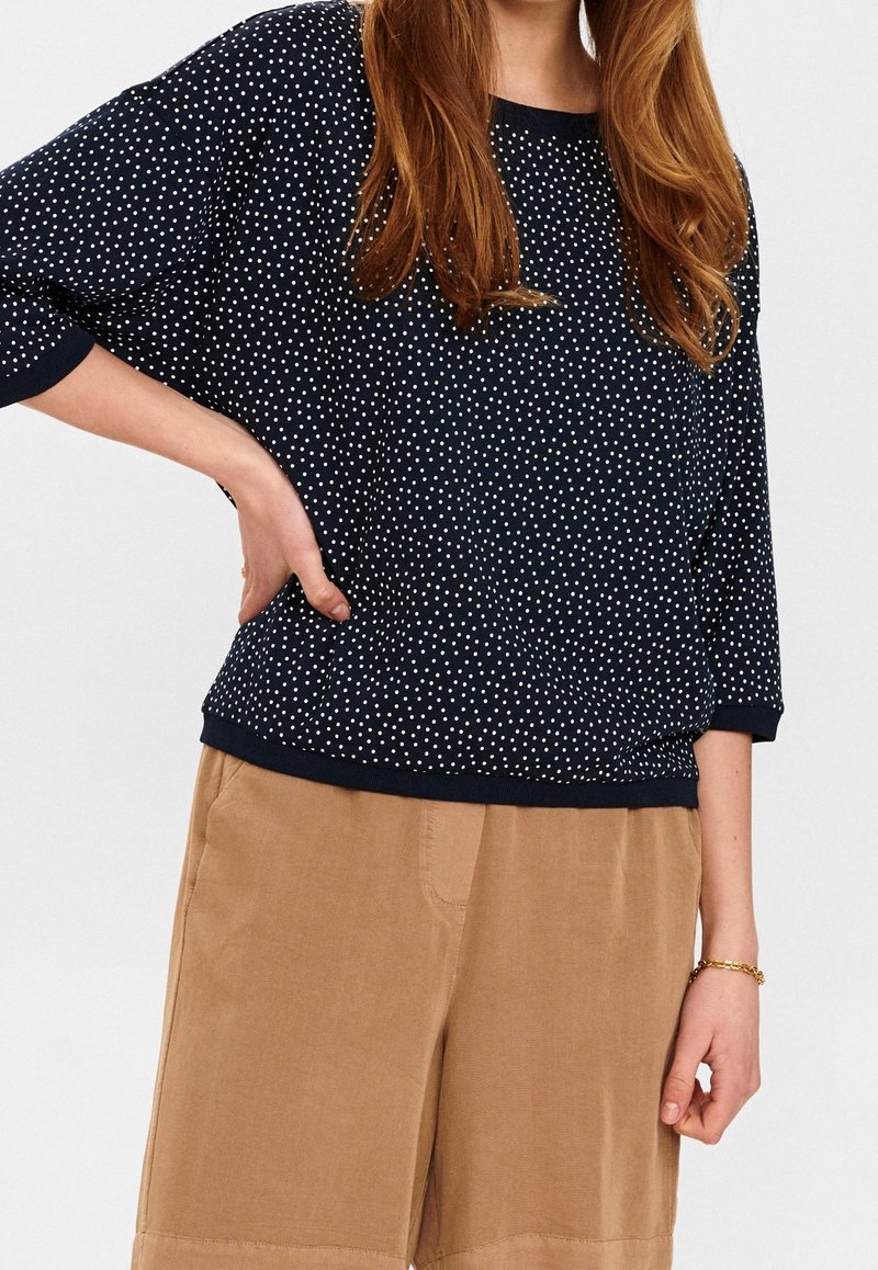 Marineblauer Pullover mit weißem Polka-Dot-Muster, Rundhalsausschnitt und Dreiviertelärmeln. Kombiniert mit beigen Shorts in entspannter Passform.