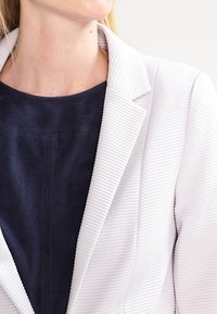 Biały żakiet w paski z wykładanym kołnierzem, noszony na ciemnoniebieskim, teksturowanym topie. Materiał wydaje się gładki z subtelnym połyskiem.