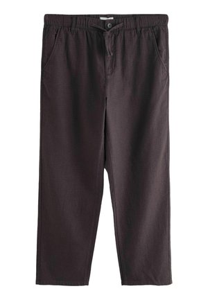 Tânăr purtând un top bej fără mâneci, cu model în dungi, pantaloni maro închis cu șnur și mocasini bej, stând pe podea de piatră lângă o ușă galbenă.