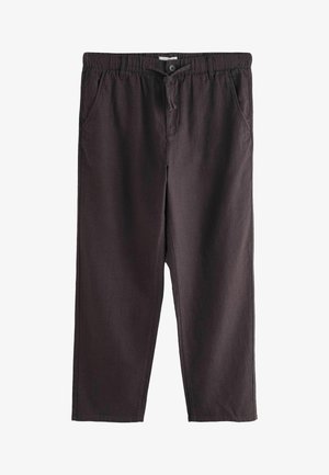 Jonge man draagt een mouwloze beige geribbelde top, donkerbruine broek met trekkoord en beige loafers, staat op een stenen vloer bij een gele deur.