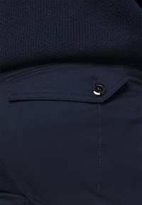 Dunkelblaue Baumwollhose mit strukturiertem Stoff, ausgestattet mit einer Fronttasche mit einem schwarzen Knopf und flachen Nähten in der Nähe des Bundes.