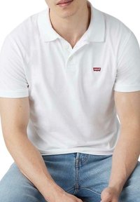 Camisa polo blanca de algodón, con un cuello clásico, mangas cortas y un pequeño logo rojo de Levi's en el lado izquierdo del pecho. Llevada con jeans azules.