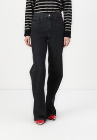 Personne portant un jean noir taille haute, un haut à manches longues rayé noir et blanc et des chaussures rouges à bouts pointus avec un motif graphique.