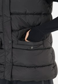 Svart pufferväst med kviltat mönster, tryckknappar och en sidoficka. Slät yta med en åtsittande passform runt torso.