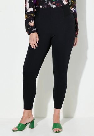 Schwarze Leggings mit glatter Textur, hoher Taille und seitlichen Taschen. Kombiniert mit grünen Absatzsandalen für einen Kontrast.