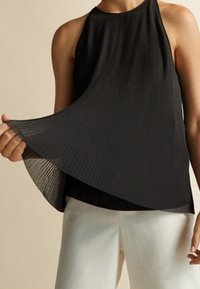 Musta pliseerattu hihaton paita, jossa on valuva muotoilu, teksturoitu kangas ja korkea kaula-aukko, käytetty valkoisten housujen kanssa.