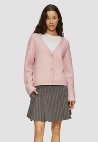Rosa gestrickte Strickjacke mit V-Ausschnitt, geripptem Muster und fünf Knöpfen. Getragen über einem weißen Hemd und kombiniert mit einem grauen gestreiften Rock.