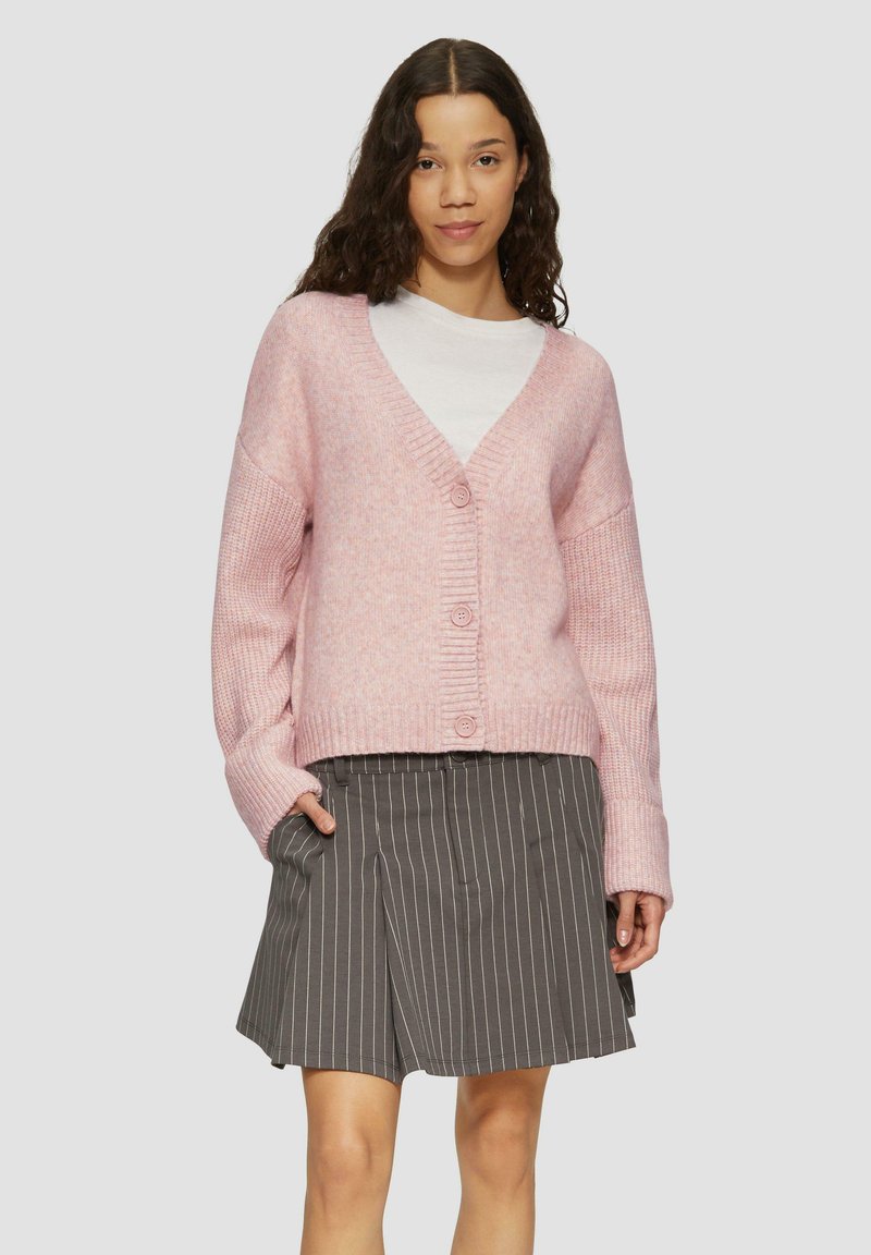 Rosa gestrickte Strickjacke mit V-Ausschnitt, geripptem Muster und fünf Knöpfen. Getragen über einem weißen Hemd und kombiniert mit einem grauen gestreiften Rock.