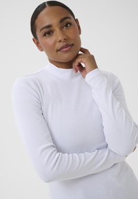 Tricou alb cu mânecă lungă și guler înalt, din material țesut cu textura netedă, purtat lejer cu brațele încrucișate.