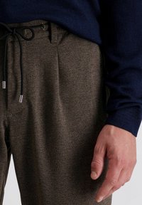Bruine getextureerde broek met een trekkoord in de taille, voorzien van metalen uiteinden. gedragen met een donkerblauwe trui, waarbij een deel van de hand zichtbaar is.