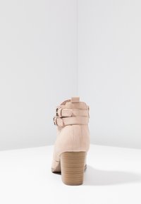 Bottine beige avec un matériau en suède texturé, deux courroies à boucle réglables, un bout arrondi et un talon en bois empilé.