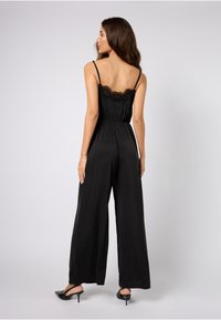 Sort satinfarge drakt med blonderkant ved halsen, justerbare stropper, elastisk midje og bred-ben design. Jevn tekstur og minimale detaljer.