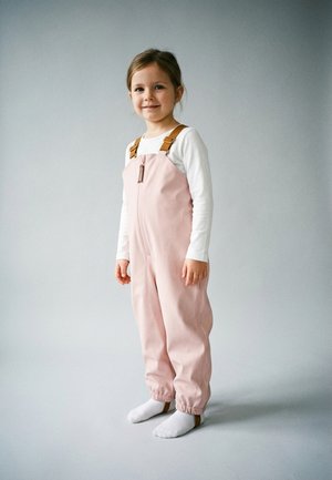 Junges Mädchen steht, trägt ein hellrosa Latzhose mit Reißverschluss und braunen Trägern über einem weißen Langarmshirt und weißen Socken, neutraler Hintergrund.