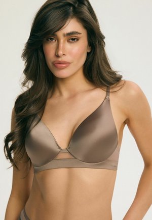Žena s dlhými tmavými vlasmi, ktorá nosí hladkú hnedú bezdrôtovú podprsenku s tenkými ramienkami na jednoduchom svetlom pozadí.