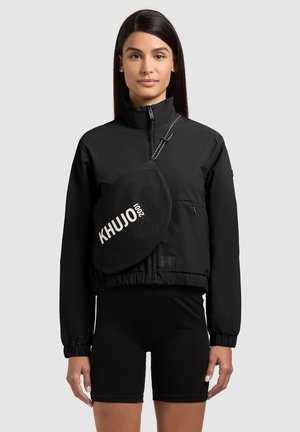 Kobieta ubrana w czarną kurtkę na zamek, czarne szorty oraz czarną torebkę na ramię z białym napisem "KHUJO 2001".