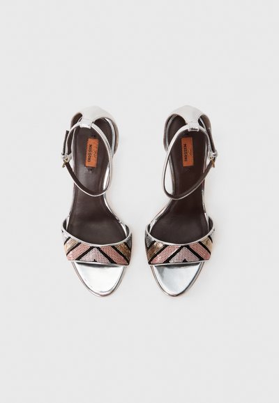 Μεταλλικά ασημένια πέδιλα με τακούνι, με σχέδιο μαύρης και χρυσής ζώνης που διαθέτει γεωμετρικά σχήματα. Ρυθμιζόμενη ζώνη στον αστράγαλο για καλύτερη εφαρμογή.