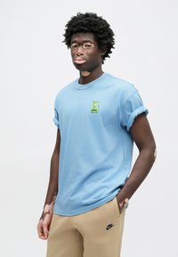 Gaiši zils kokvilnas T-krekls ar uzlocītām īsām piedurknēm, uz kreisā krūškurvja zaļš karikatūras uzraksts, valkāts kopā ar bēšām sporta biksēm.