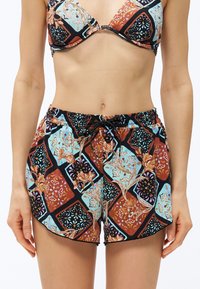 Γυναίκα που φοράει τοπ μπικίνι με σχέδια και αντίστοιχα σορτς με κορδόνι, με γεωμετρικό και λουλουδάτο σχέδιο σε μαύρο, μπλε και σκουριά χρώματα.