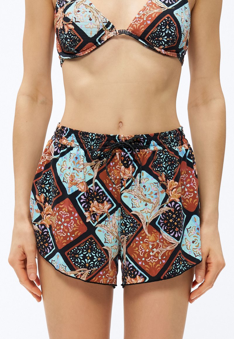 Γυναίκα που φοράει τοπ μπικίνι με σχέδια και αντίστοιχα σορτς με κορδόνι, με γεωμετρικό και λουλουδάτο σχέδιο σε μαύρο, μπλε και σκουριά χρώματα.