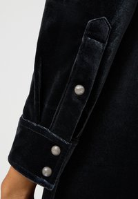 Indumento in velluto nero con un polsino caratterizzato da due grandi bottoni tondi e lucidi e un design strutturato lungo il bordo della manica.