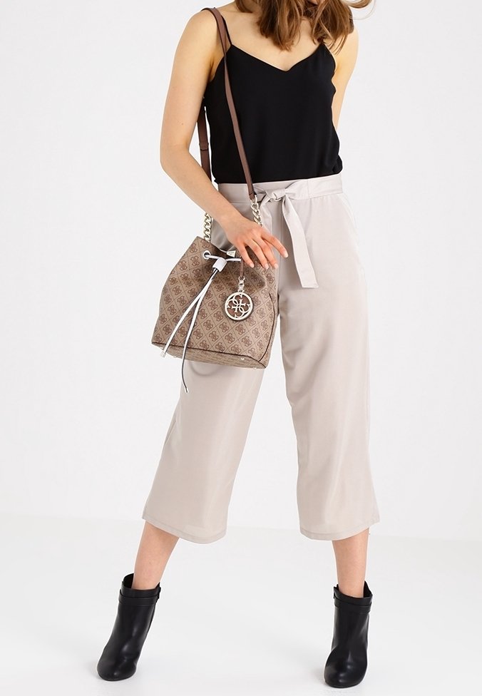 Femme porte un pantalon beige à jambes larges, un haut noir sans manches, des bottines noires, tenant un sac seau beige à motif avec cordon de serrage et bandoulières en chaîne.