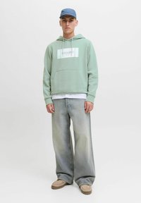 Jonge man in een lichtgroene hoodie, vervaagde wijde jeans, beige schoenen en een blauwe pet, staand tegen een effen achtergrond.