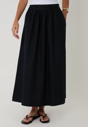 Jupe longue noire à taille froncée portée avec des tongs noires et un haut blanc ; la main gauche du modèle dans la poche de la jupe et la main droite détendue.