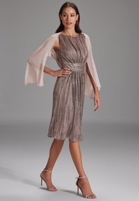 Robe mi-longue en argent métallique avec des rayures roses, taille cintrée et manches fluides en dentelle. Associée à des talons métalliques à lanières.