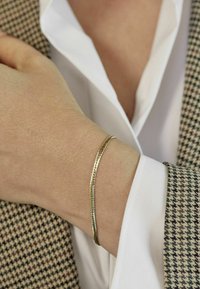 Tunn guld-färgad armband med en texturerad mönster, bärs på en handled, parat med en vit blus och en hounds-tooth-mönstrad kavaj.