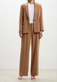 Ensemble costume beige comprenant un blazer ajusté avec des revers à crans et un pantalon à jambes larges. Les matériaux semblent lisses, avec un léger éclat.