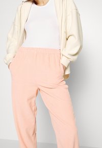 Měkké, světle růžové ribbed kalhoty s elastickým pasem a postranními kapsami, v kombinaci s bílým upnutým topem a krémovou mikinou na zip.