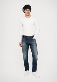 Bílá košile s dlouhým rukávem a V výstřihem, tmavě modré džíny, černý pásek a bílé tenisky. Neformální outfit s úzkou siluetou a mírným odřením na džínách.