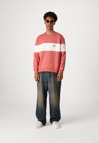 Garās piedurknes korallveida sporta krekls ar baltu horizontālu svītru un logotipu, apvienots ar brīvām tumšām džinsām un baltām kedām. Gluda auduma tekstūra.