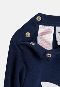 Cămașă din bumbac albastru marin cu butoane aurii la gât, având un logo imprimat și o etichetă de întreținere pe interior.