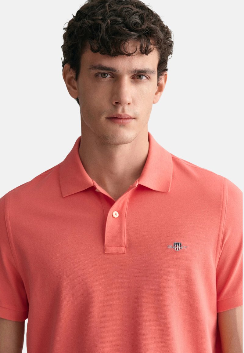 Koralfarbenes Poloshirt aus strukturiertem Stoff. Verfügt über einen klassischen Kragen, eine Zwei-Knopf-Leiste und ein kleines gesticktes Logo auf der Brust.