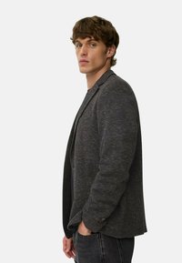 Grafitgrauer Blazer mit strukturiertem Finish, Ein-Knopf-Verschluss und spitzen Revers, kombiniert mit einem schwarzen Hemd und Jeans.