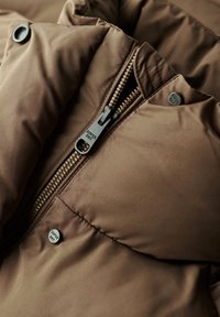 Bruine pufferjasstof met een glad oppervlak, voorzien van een zwarte ritssluiting en een gebrande knoop. De binnenkant toont details van zachte isolatie.