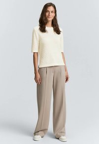 Ljus krämfärgad tröja med struktur och korta ärmar, kombinerad med löst sittande beige byxor och vita sneakers, stående mot en ljus bakgrund.