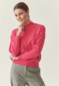 Pull fuchsia avec un col haut, des poignets et un ourlet côtelés, associé à un pantalon gris clair. Texture lisse et coupe décontractée.