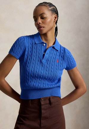 Γυναίκα με πλεξούδες, φορέι μπλε πόλο μπλούζα με κοντά μανίκια και πλεκτό σχέδιο, και καφέ παντελόνι, ποζάρει με τα χέρια στη μέση μπροστά σε απλό φόντο.