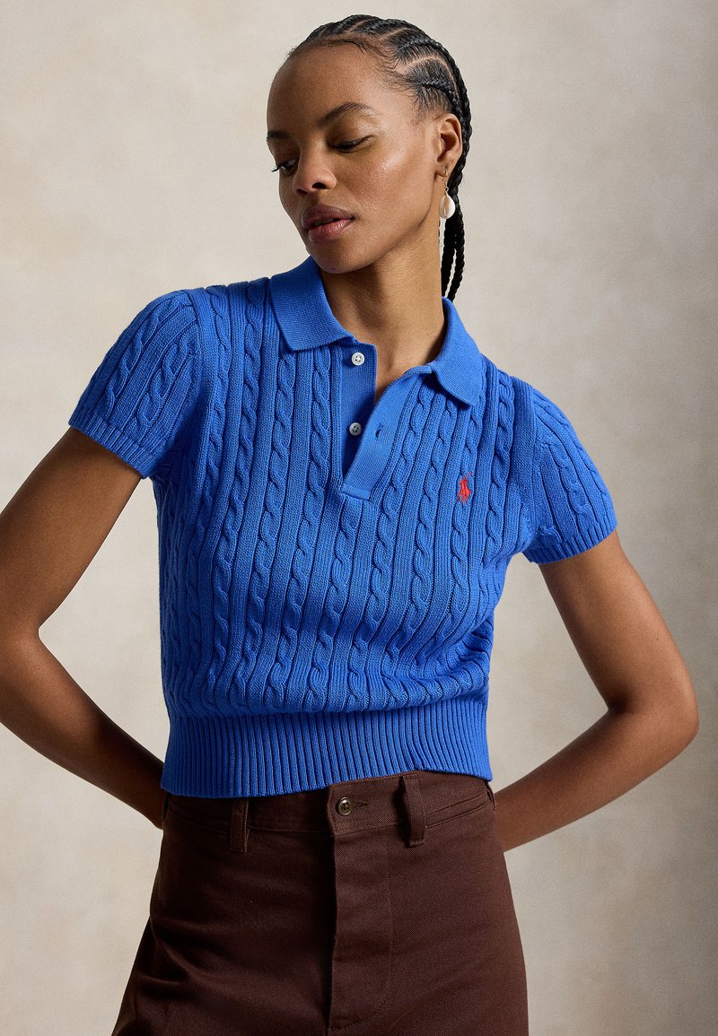 Femme aux cheveux tressés portant un polo bleu à manches courtes en maille torsadée et un pantalon marron, posant les mains sur les hanches devant un fond uni.