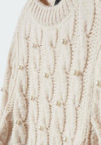 Pull en tricot crème avec un col montant côtelé, un motif torsadé texturé et des ornements décoratifs en perles partout.