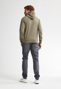 Felpa verde oliva con cappuccio regolabile, polsini e vita a costine, abbinata a jeans in denim grigi e sneaker bianche. Vista posteriore mostrata.